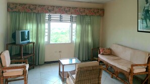 A living room with a television on a wooden stand, a wicker sofa, a glass coffee table, and a view of greenery outside through the window. - Symphony Resort (Discovery Bay)