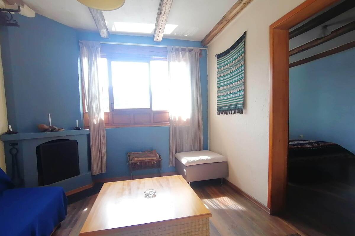 A room with a wooden table, a blue sofa, a small ottoman, a fireplace, and a window with curtains.