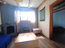 Una habitación con una mesa de madera, un sofá azul, un pequeño reposapiés, una chimenea y una ventana con cortinas.