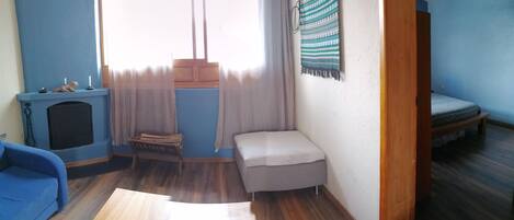 Una habitación con pared azul, un banco pequeño de madera, un sofá gris, una ventana con cortinas traslúcidas y una cama con colchón gris.