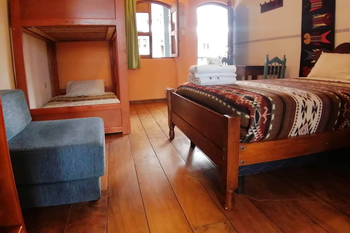 A room with a bunk bed, a single bed with a patterned blanket, a chair, and a window with curtains.