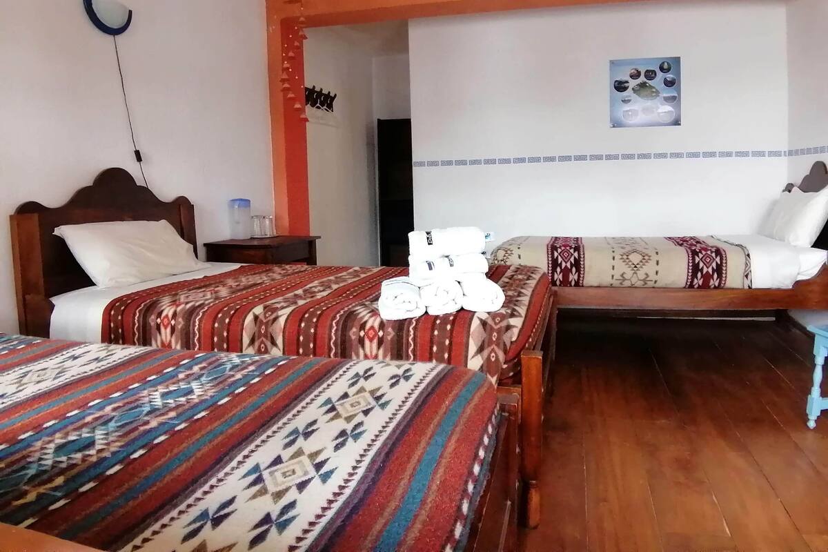 A hotel room with two beds, each with patterned bedding, a wooden headboard, a bedside table, and a wall-mounted light fixture.