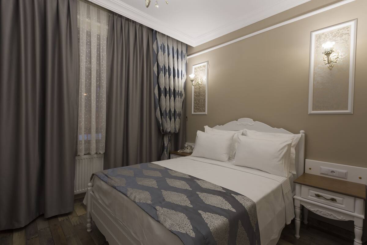 Una habitación de hotel con cama, mesita de noche, lámpara y ventana con cortinas.