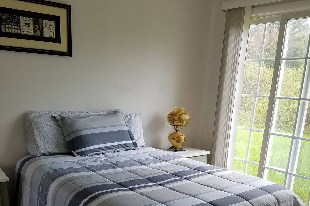 Un dormitorio con cama, una mesita de noche, un cuadro enmarcado y una ventana con vista a follaje.