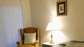 A hotel room with a large bed, a chair, a nightstand with a lamp, and a window with blinds. - Becket Motel (Becket)