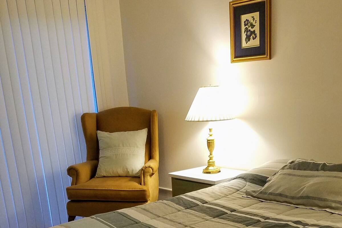 Habitación de hotel con una cama grande, una silla, una mesita de noche con lámpara y una ventana con persianas.