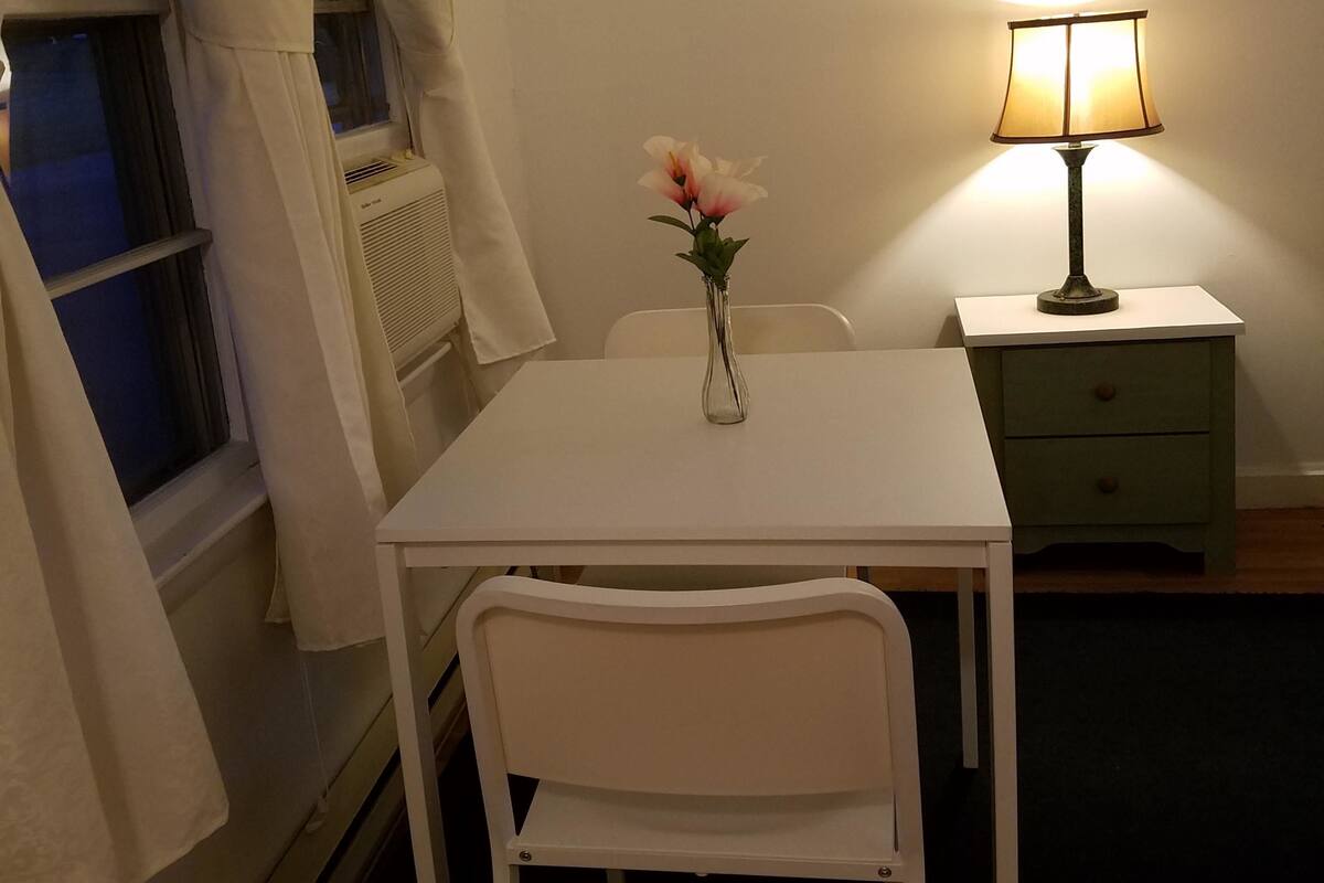 Una habitación pequeña y limpia con una mesa blanca, una silla, una mesita de noche con lámpara y una ventana con cortinas.