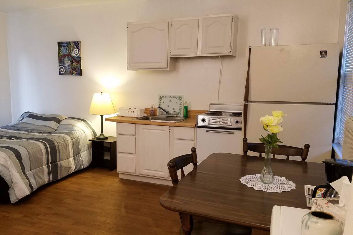Habitación compacta con cama, kitchenette pequeño, mesa de comedor con sillas y ventana con persianas.