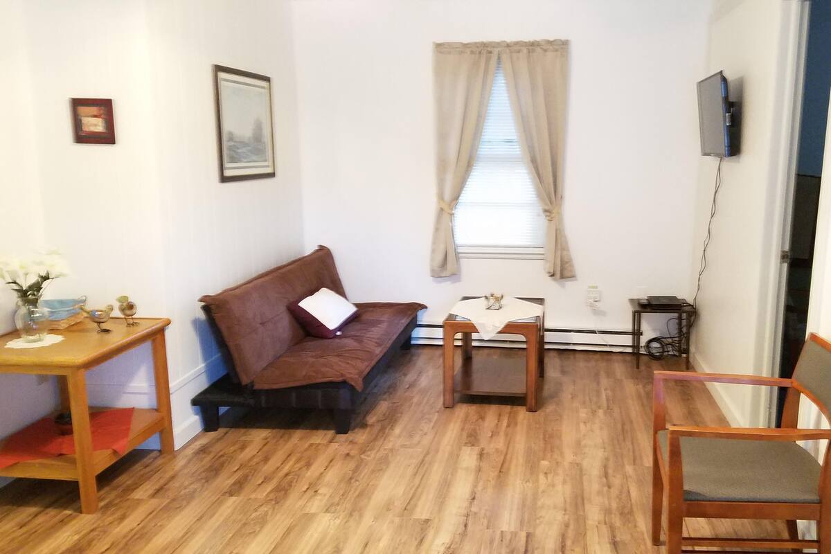 Una habitación pequeña y amoblada, con piso de madera, un sofá marrón, una silla de madera, una mesita con un jarrón y un televisor montado en la pared.