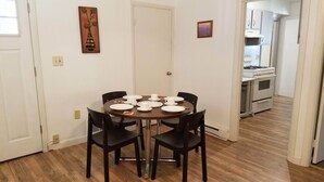 A dining area with a round table set for four, surrounded by dark chairs. A white door is visible in the background, leading to a kitchen area with white appliances. - Becket Motel (Becket)