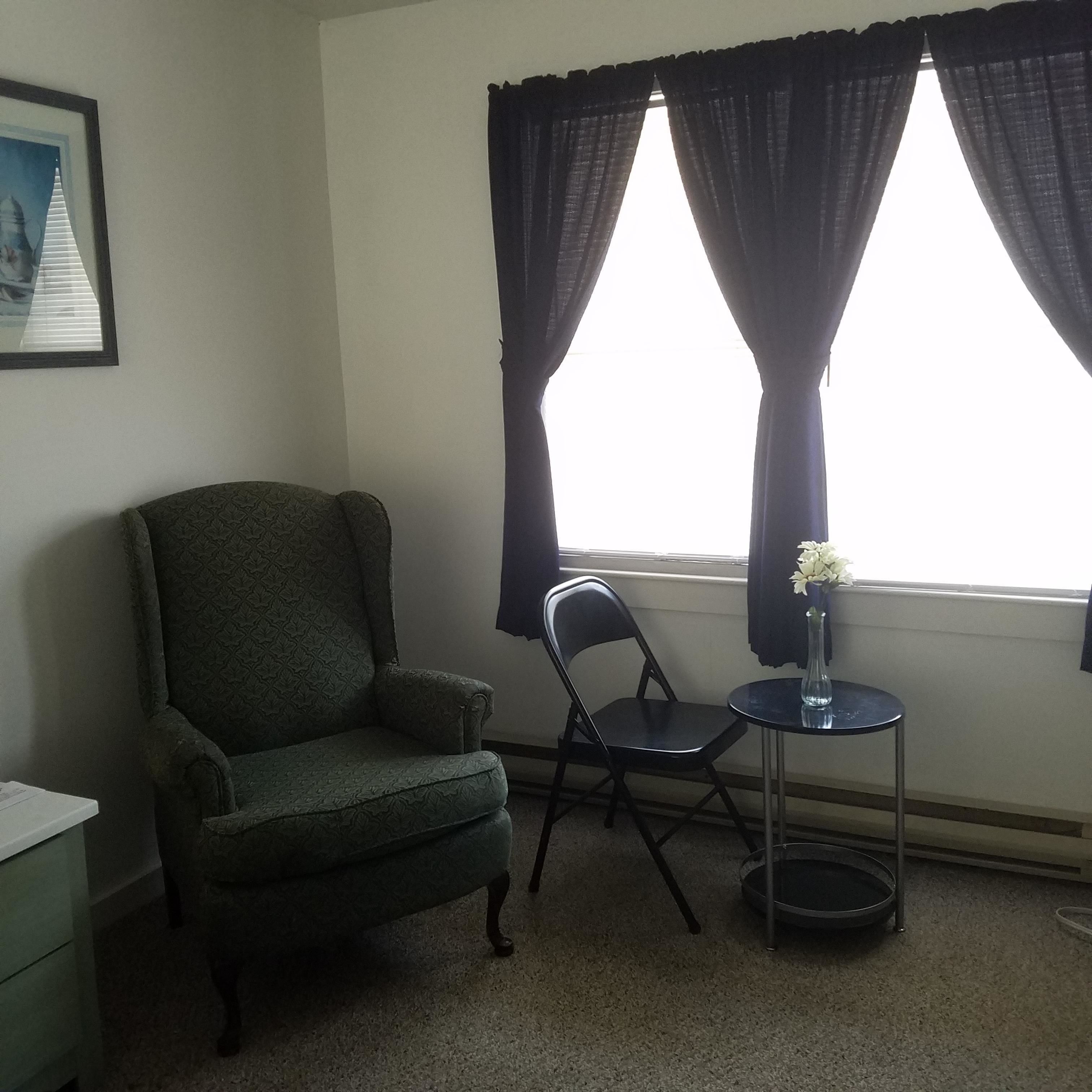 Um quarto com uma poltrona verde, uma cadeira preta, uma mesa redonda pequena com um vaso de flores e uma janela com cortinas escuras.