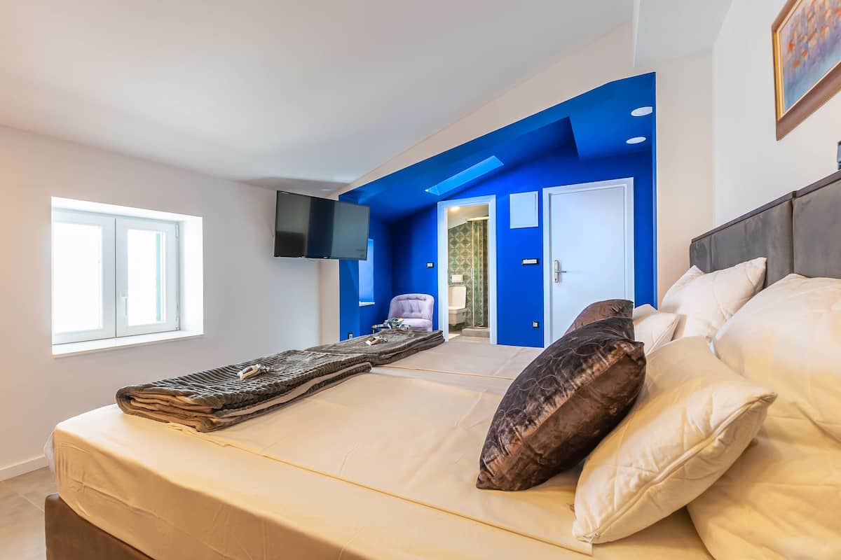 Quarto com cama, almofadas, televisão, janela e uma parede azul de destaque.
