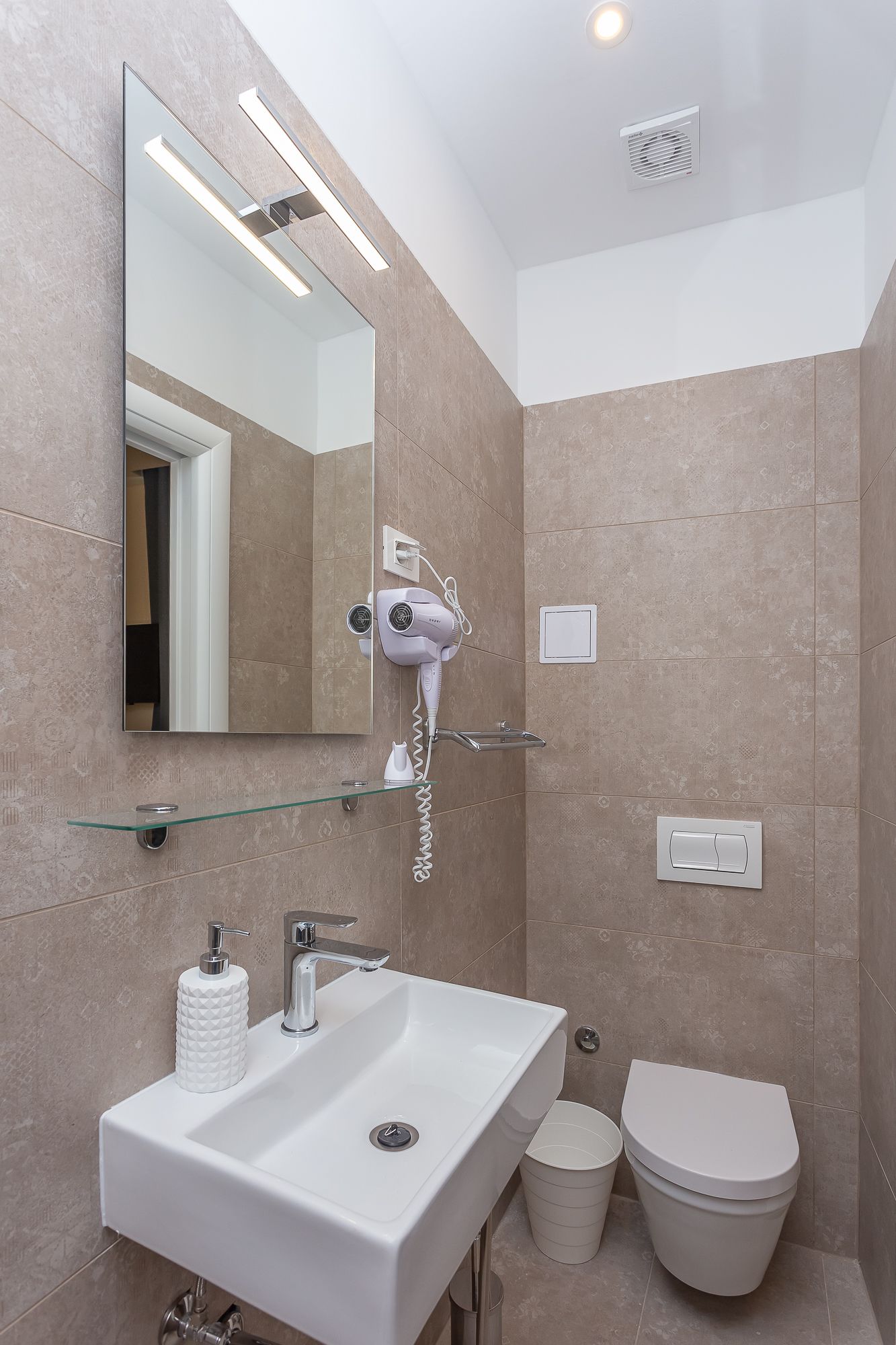 A modern bathroom with a large mirror, a hairdryer mounted on the wall, a white sink, and a toilet.