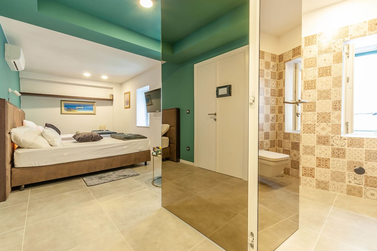 Quarto com cama, mesa de cabeceira e apoio para objetos. Possui banheiro com vaso sanitário, pia e chuveiro. O teto é verde-água e as paredes são bege.