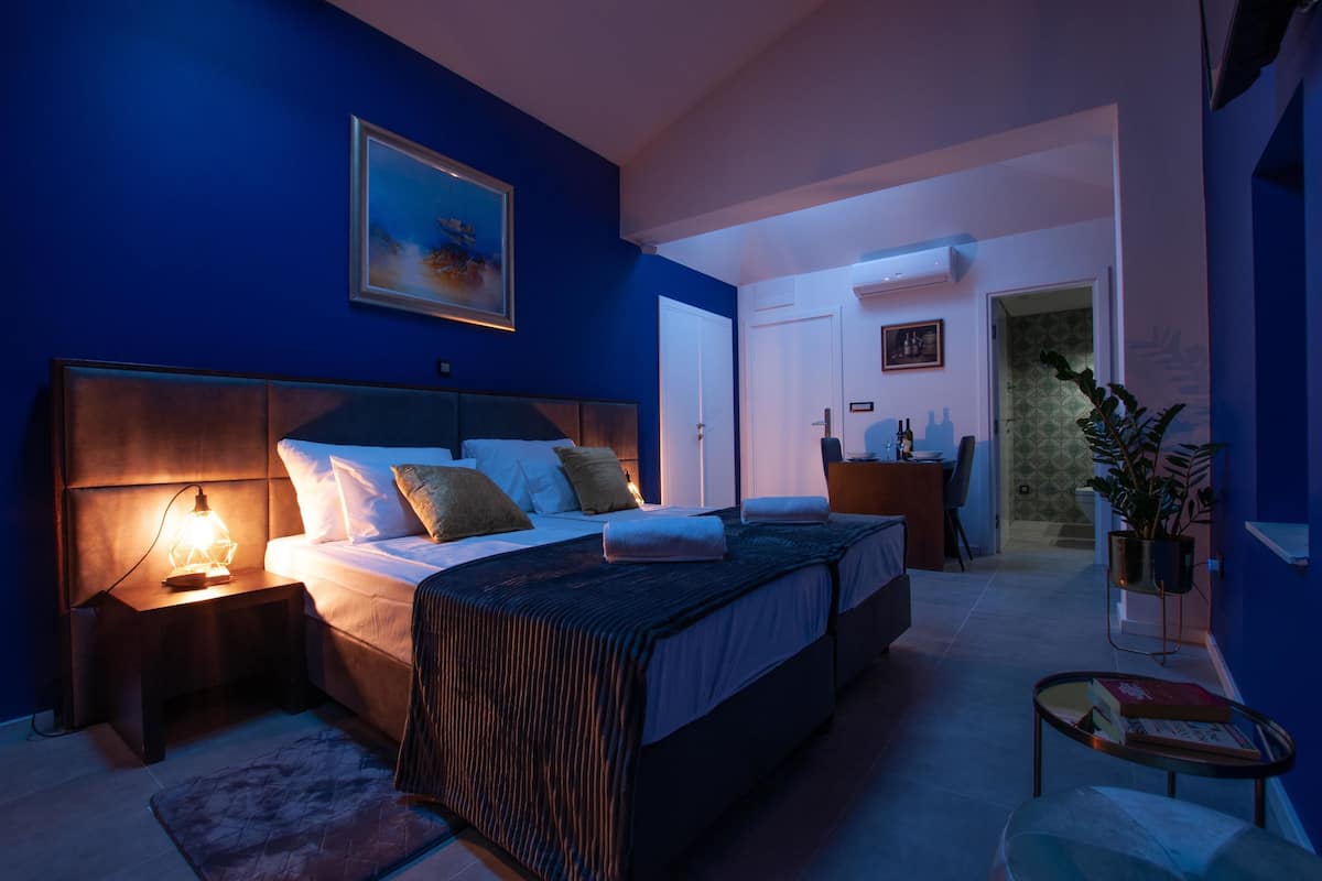 Quarto com uma cama grande, mesas de cabeceira, um criado-mudo, um quadro pendurado na parede e uma porta que dá para outro cômodo.
