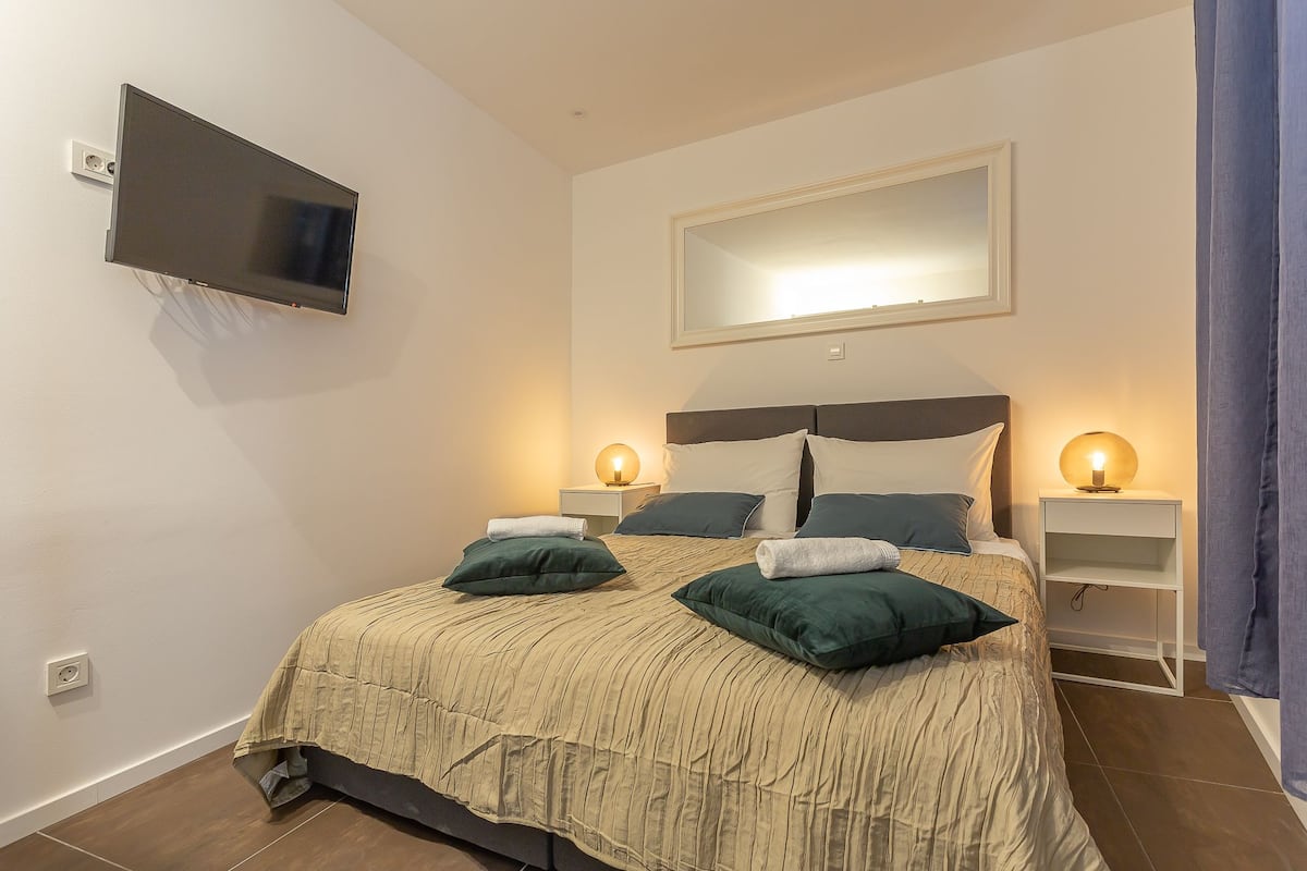 Quarto bem arrumado com uma cama, duas mesas de cabeceira, uma TV fixada na parede e um espelho sobre a cama.
