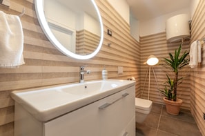 A modern bathroom with a white sink, mirror, and toilet. There is a potted plant and a wall-mounted light fixture.