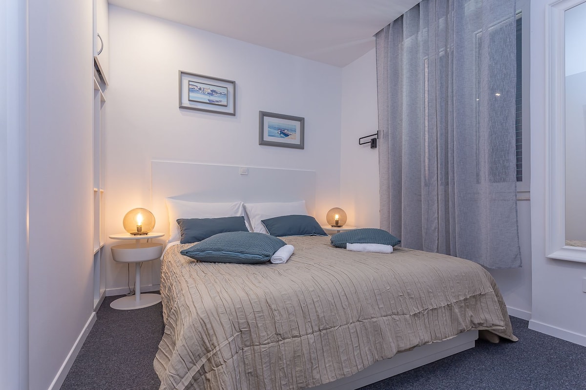 Um quarto com uma cama, duas mesas de cabeceira, uma janela com cortinas e duas imagens emolduradas na parede.