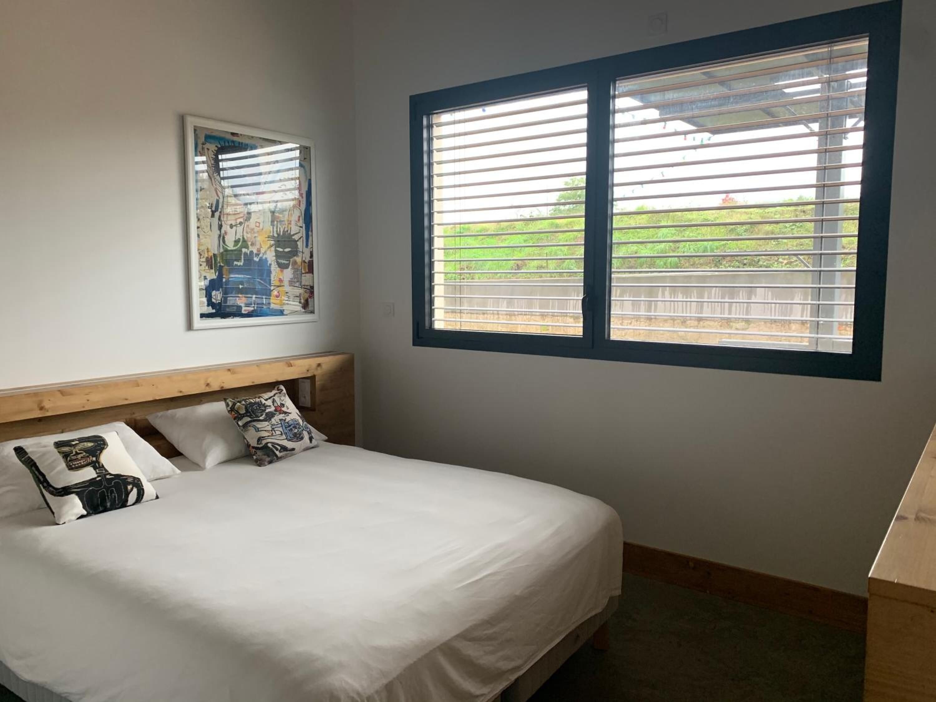 Un dormitorio con una cama, cabecera de madera, una ventana con persianas y un cuadro en la pared.