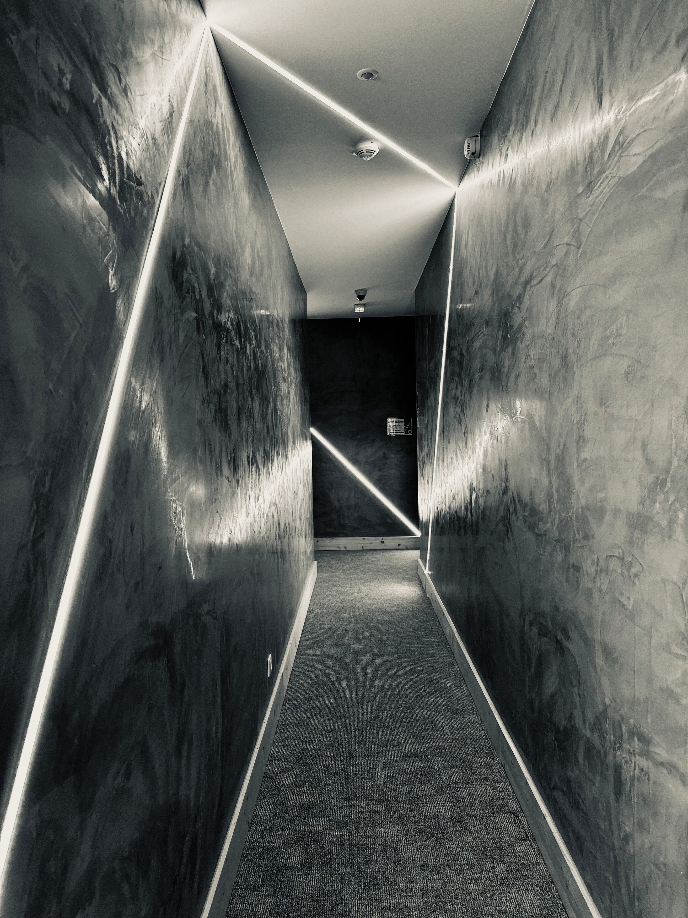 A narrow, modern hallway with reflective walls and a carpeted floor, leading to a dark room with a door at the end.