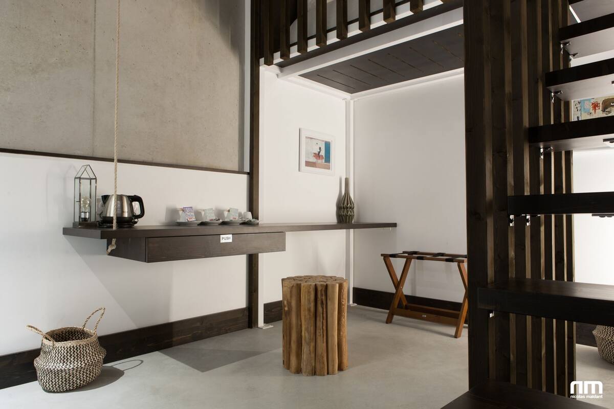 A modern interior with a wooden bench, a small wooden stool, a hanging light fixture, and a shelf with a kettle and cups.