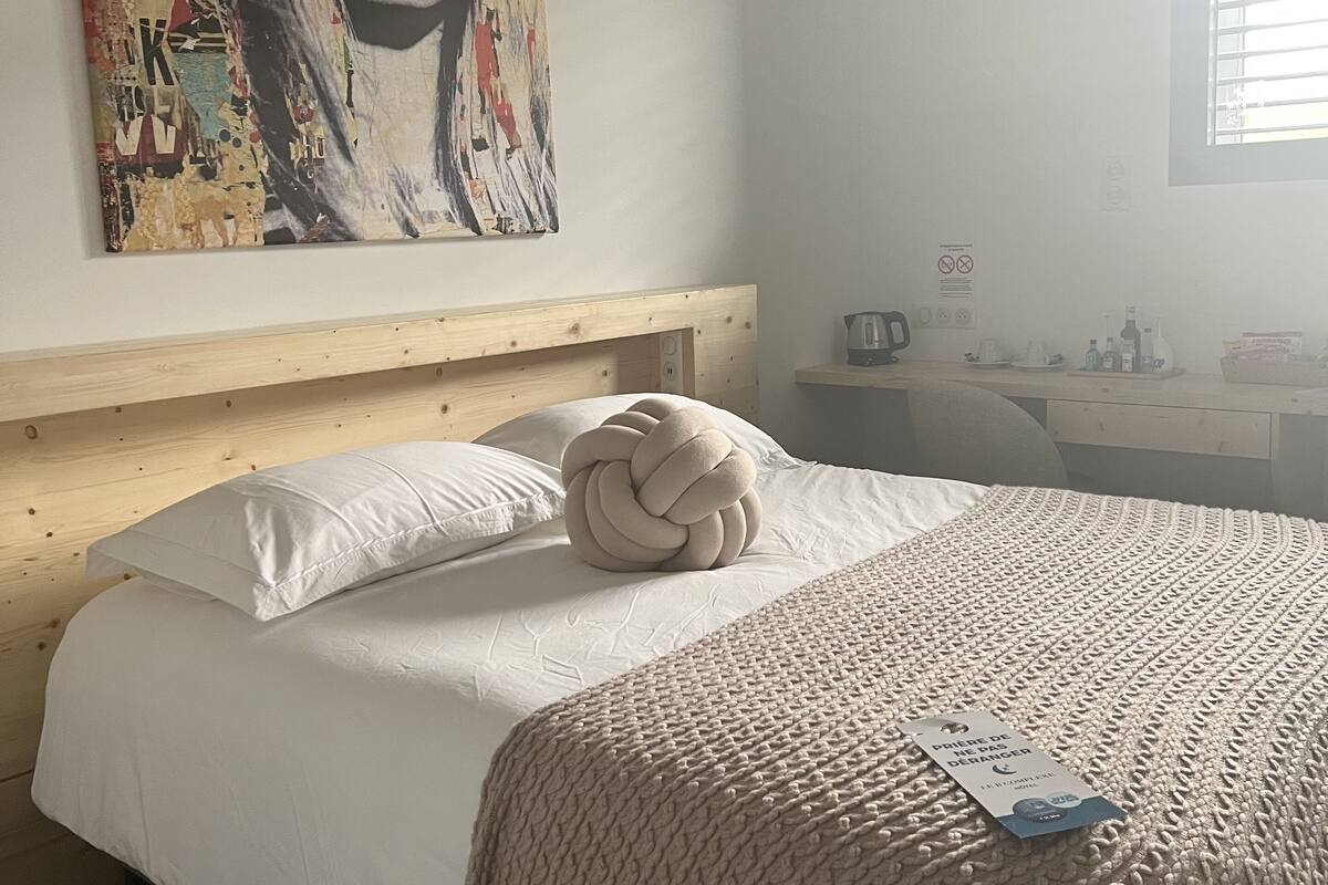 A neatly made bed with a white comforter and a knitted throw blanket, a wooden headboard, and a large abstract painting on the wall.