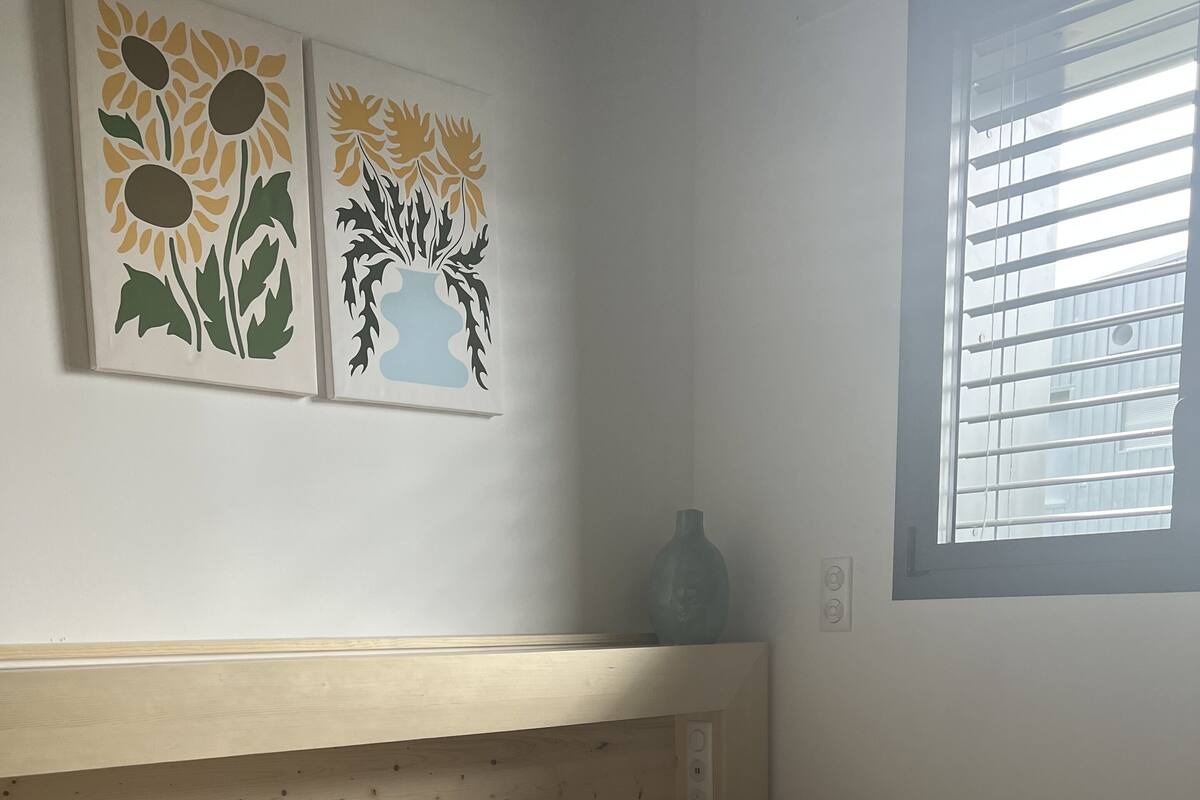 A bedroom with a wooden headboard, white bedding, a yellow flower-shaped pillow, and two wall art pieces depicting sunflowers.