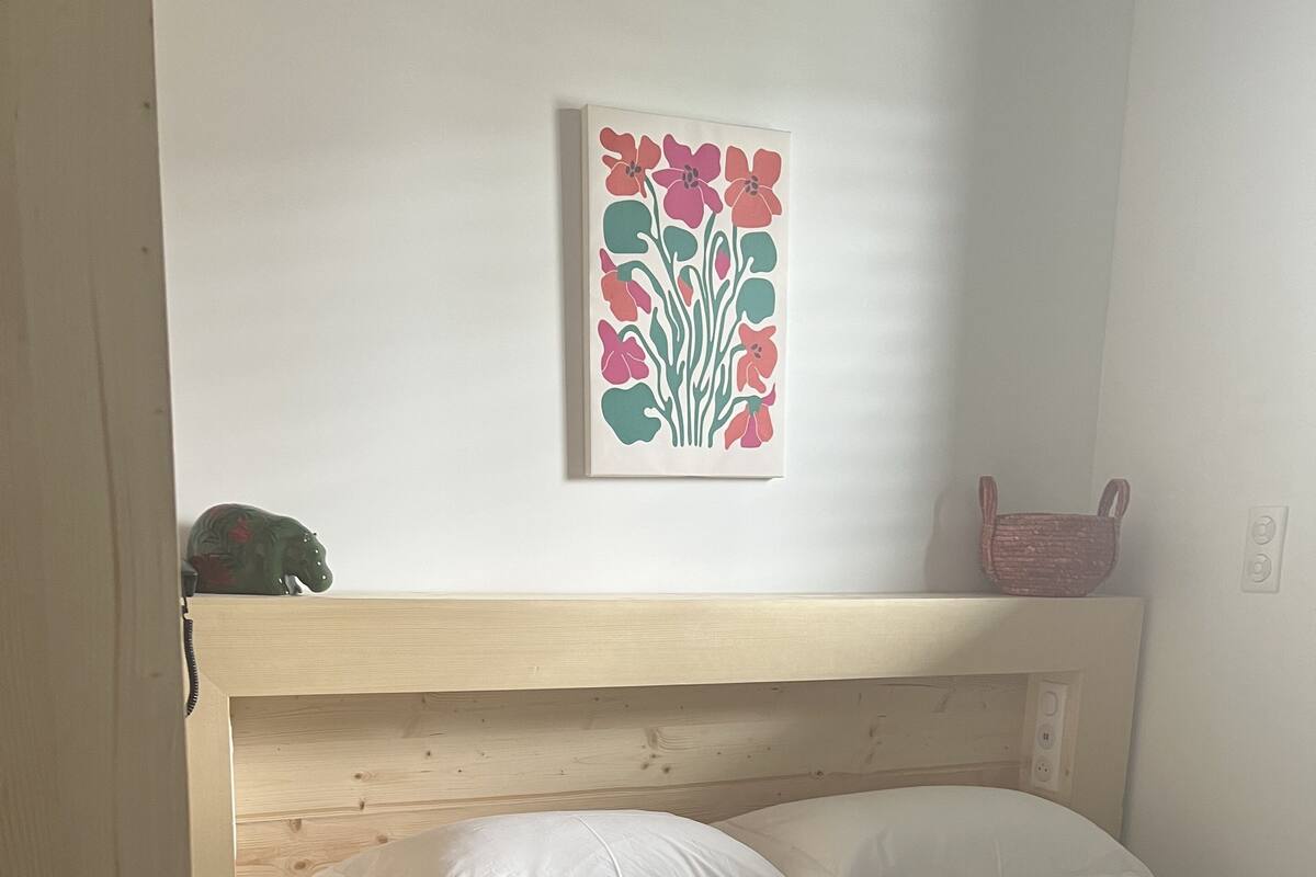 A single bed with white bedding and a green quilt, a wooden headboard, a wall-mounted artwork of flowers, and a small basket on the wall.