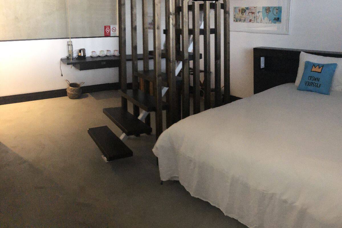 A hotel room with a bed, a wooden partition, a shelf with cups, and a small basket on the floor.