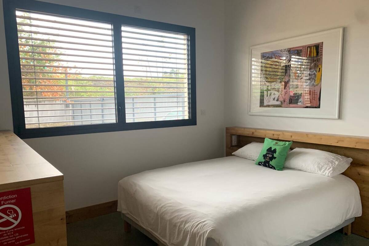 A bedroom with a bed, a window with blinds, a wooden headboard, a framed picture on the wall, and a sign on the wall.