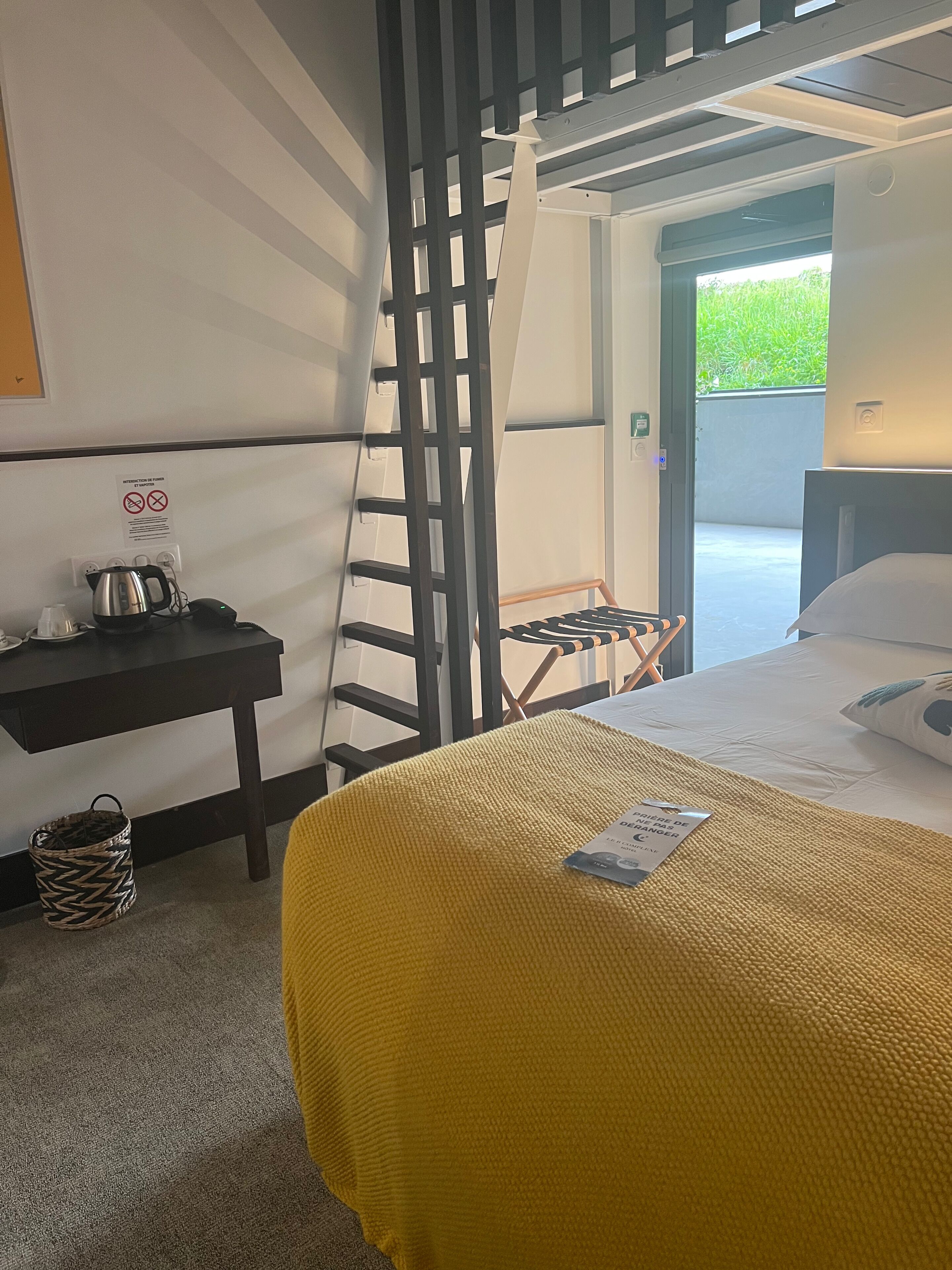A hotel room with a bed, a desk with a kettle, a chair, and a view of greenery outside.