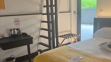A hotel room with a bed, a desk with a kettle, a chair, and a view of greenery outside.