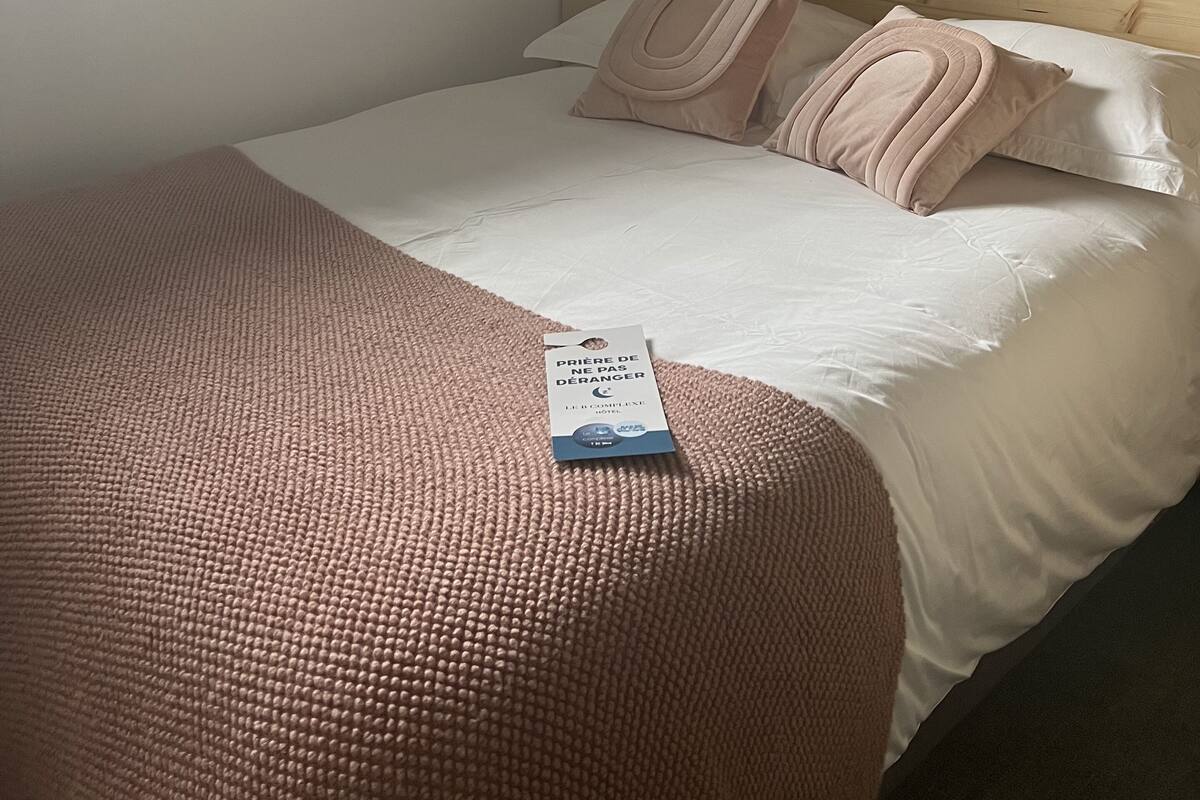 A neatly made bed with a pink blanket and pillows, a wooden headboard, a window with blinds, and a wall-mounted artwork.