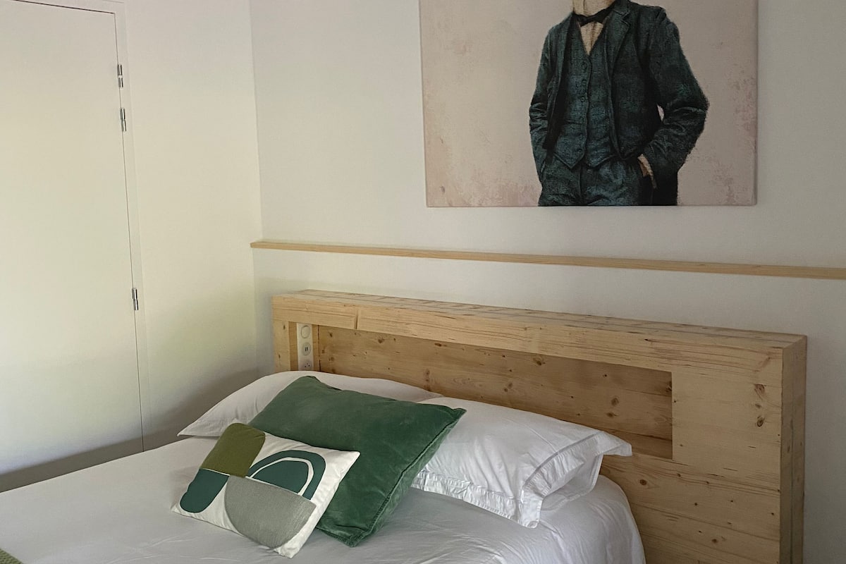 A neatly made bed with white linens and green pillows, a wooden headboard, and a painting of a giraffe on the wall.