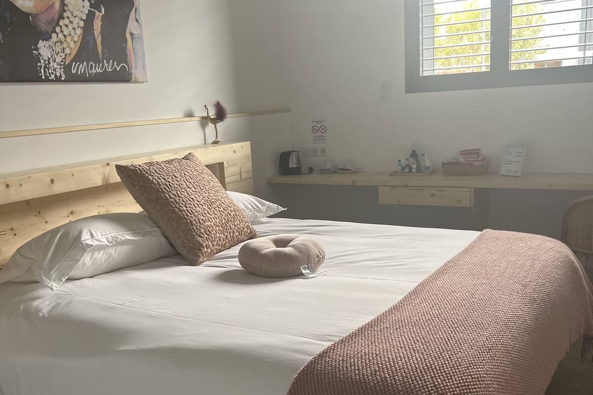 A neatly made bed with a white comforter and two pillows, a pink throw blanket, a wooden headboard, a bedside table with a lamp, and a window with blinds.