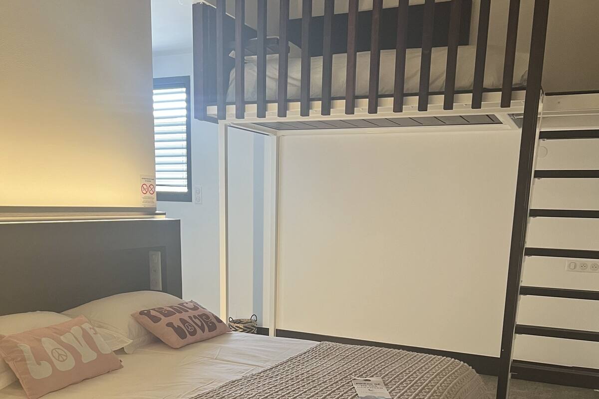 A bedroom with a bed, a headboard, a bedside table, a wall-mounted shelf, a window with blinds, and a staircase.