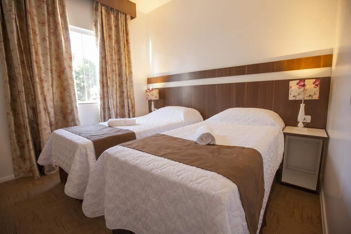 Habitación de hotel con dos camas, piso de madera, una ventana con cortinas y una mesita de noche con lámpara.