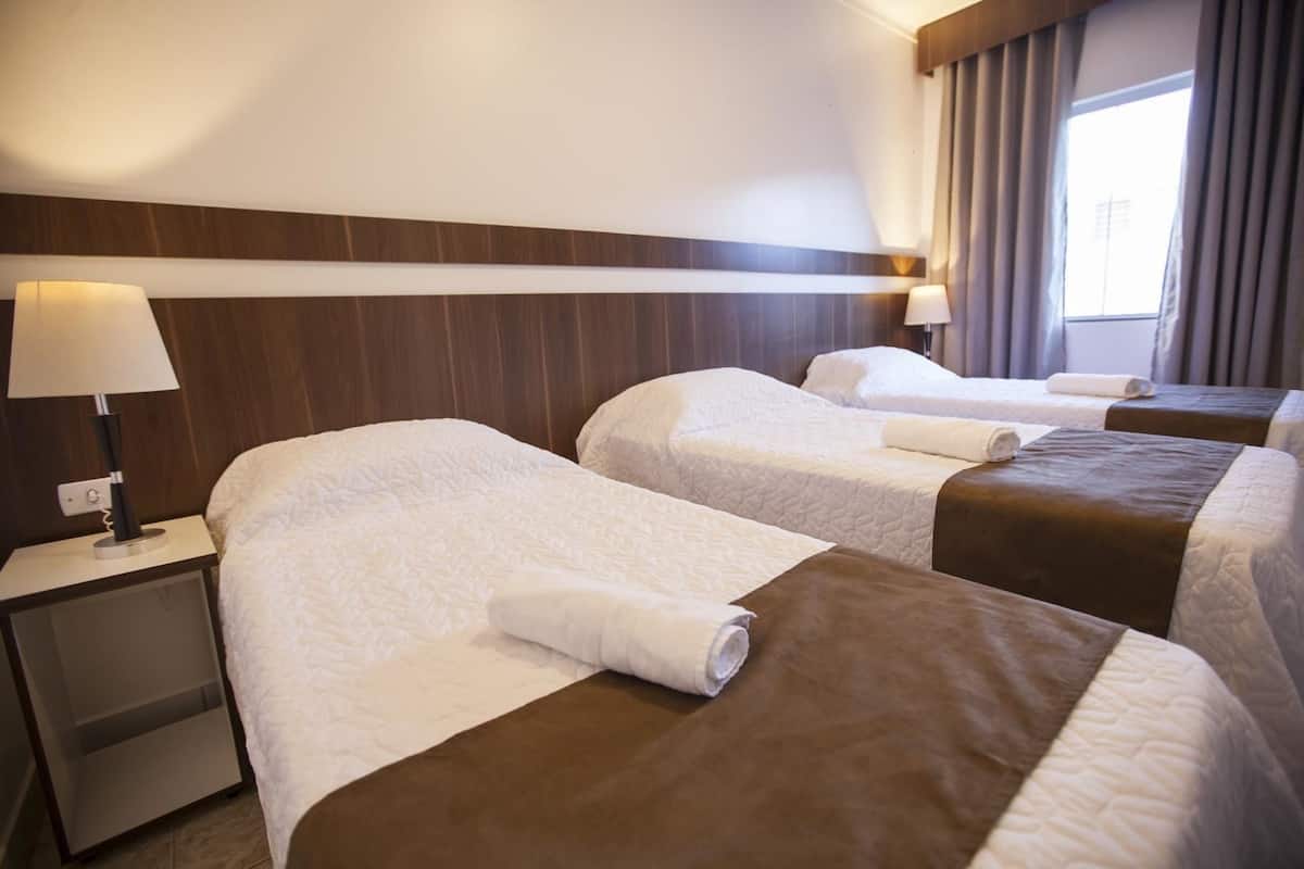 Habitación de hotel con tres camas, cada una con cubrecama blanco y marrón, y una mesita de noche con lámpara.