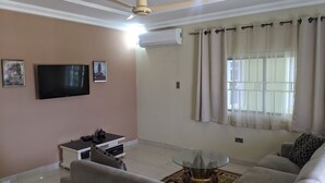 A living room with a ceiling fan, a flat-screen TV, a sofa, a coffee table, and a window with curtains. - Jacksonfields Apartments (Accra)