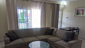 A living room with a beige sectional sofa, two patterned throw pillows, a glass coffee table, and a patterned rug. - Jacksonfields Apartments (Accra)