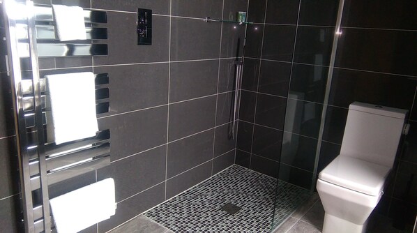 A modern bathroom with dark tiles, a glass shower enclosure, a toilet, and a metal vent. - The Castle Apartments (Castletown)