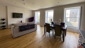 A spacious living room with a sofa, dining table, and chairs. There is a TV mounted on the wall and large windows providing natural light. - The Castle Apartments (Castletown)