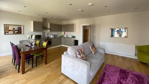 A modern living room with a grey sofa, dining table, and chairs. A kitchen is visible in the background with grey cabinets and a white countertop. - The Castle Apartments (Castletown)
