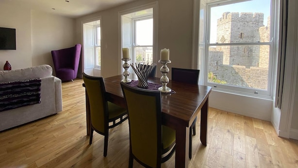 A dining area with a wooden table and chairs, a purple armchair, and a view of a stone building through large windows. - The Castle Apartments (Castletown)