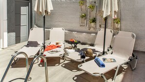 Un espacio al aire libre iluminado por el sol con sillones de descanso, sombrillas y una pequeña mesa con una cesta y una botella.