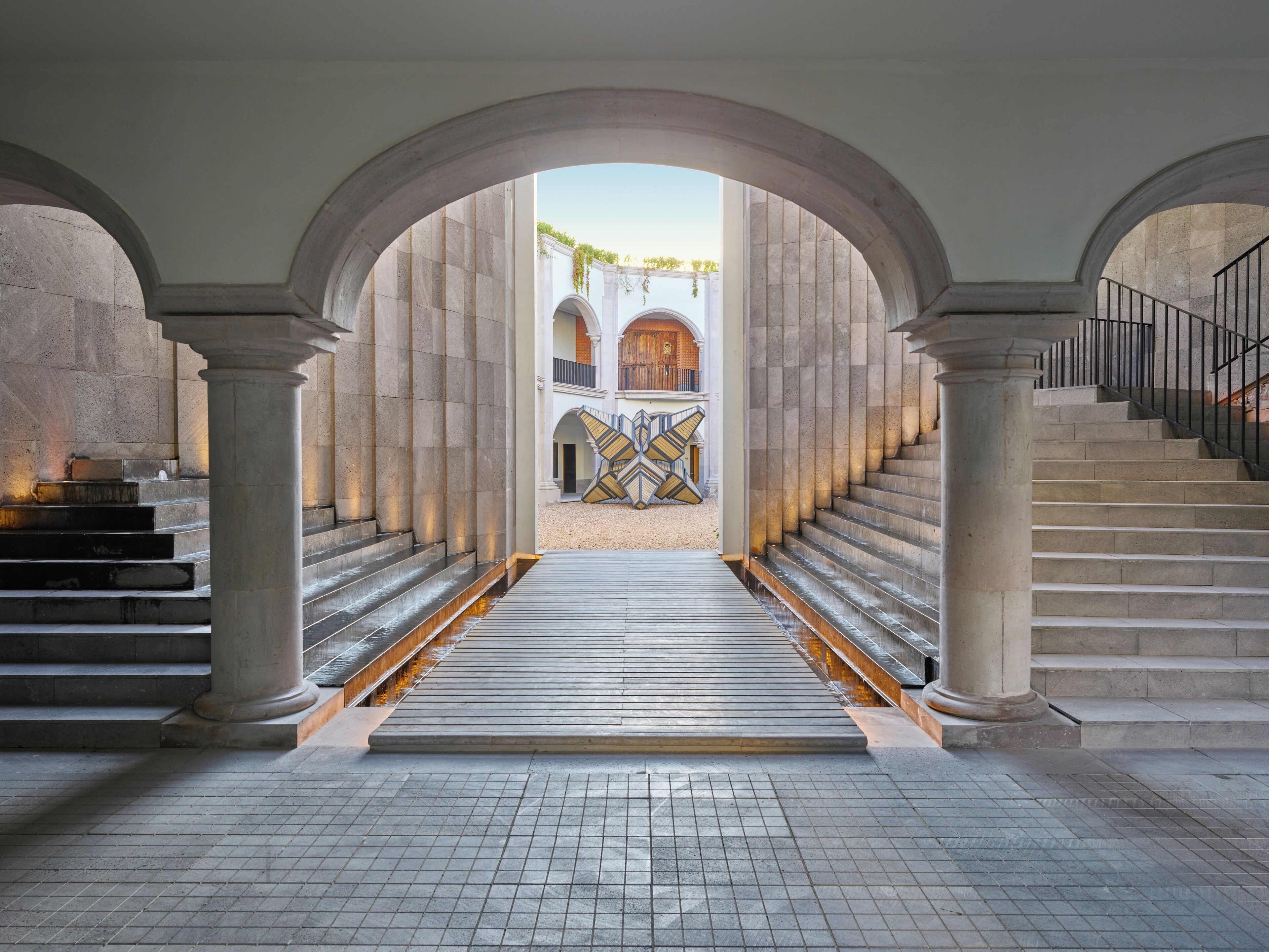 Un amplio vestíbulo con una escalera central flanqueada por imponentes columnas de piedra, que conduce a un edificio con arcos.
