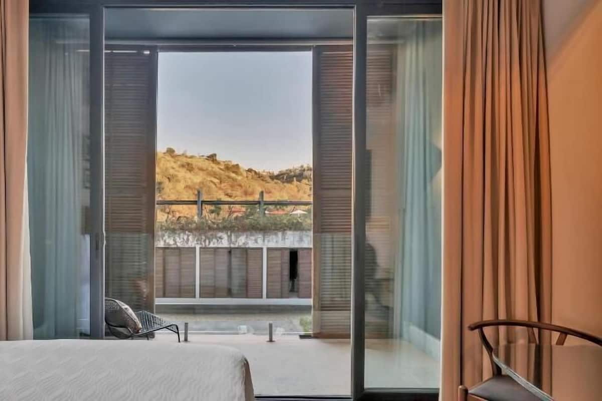 Quarto com cama, uma cadeira e varanda com vista para uma encosta.