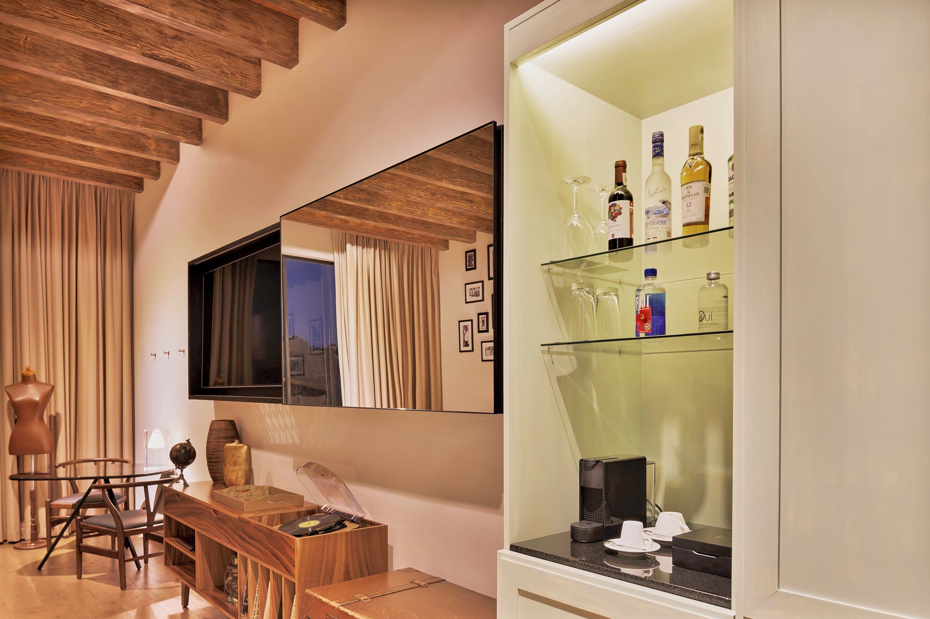 Una habitación moderna con techo de madera, un espejo grande, un aparador de vidrio con botellas y un escritorio de madera con sillas.