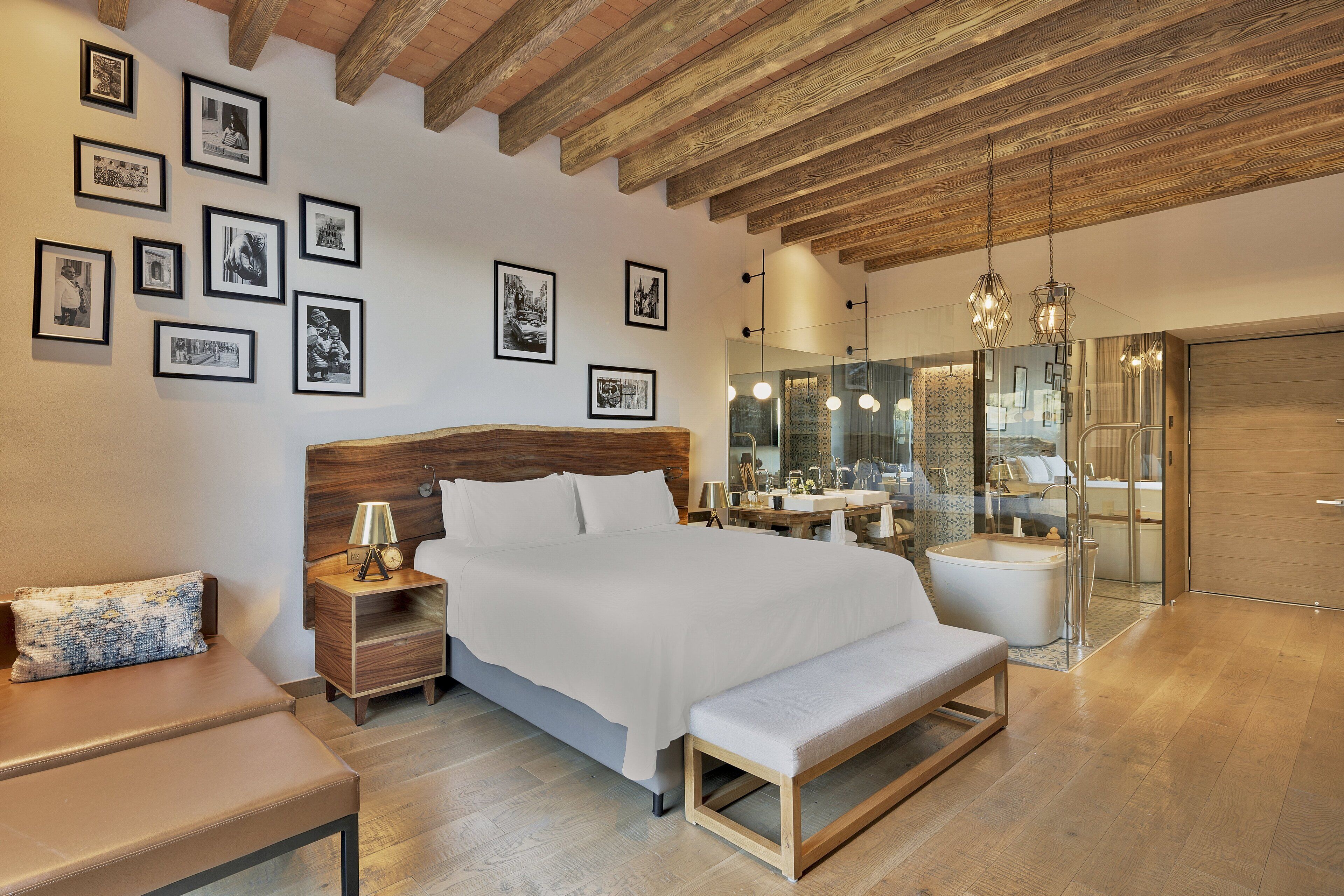 A modern bedroom with a large bed, wooden beams, a bench, bedside tables, and framed pictures on the wall.