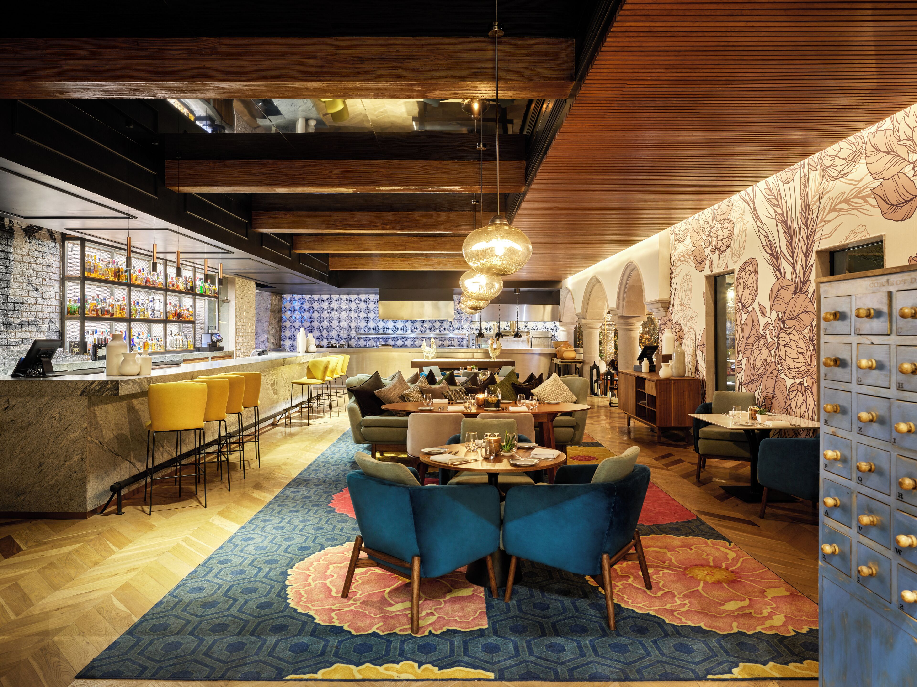 A modern bar area with a wooden ceiling, a bar counter with a variety of bottles, and seating arrangements including bar stools and cushioned chairs.
